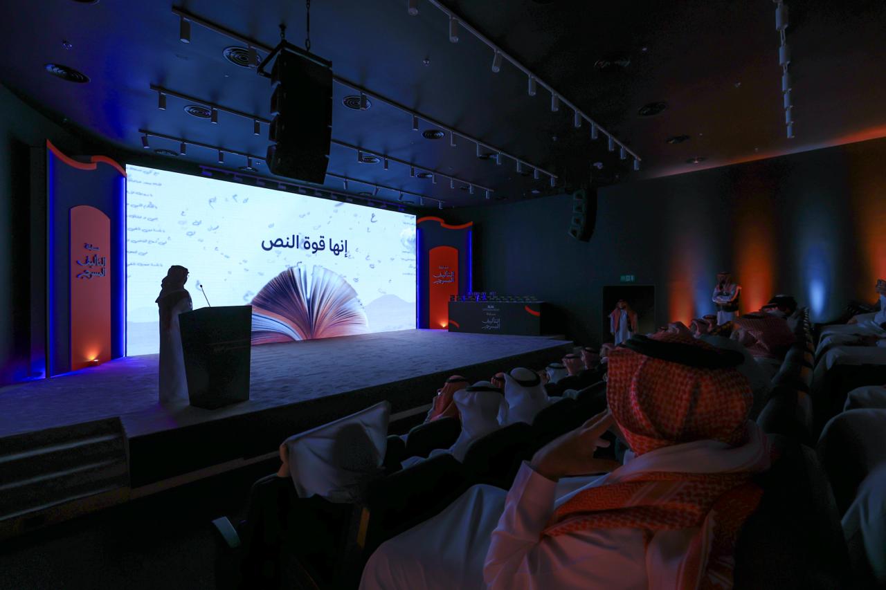 قاعة معتمة في حفل مسابقة التأليف المسرحي: متحدّث على منصّة إلى جانب شاشة كبيرة تعرض كتابًا مفتوحًا وعبارة «إنها قوة النص»، أمام جمهور يملأ المقاعد.
